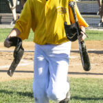 Batboy, Mason, trots back to the dugout from homeplate carrying a bat and an elbow guard (versus Novato Knicks, Sunday, July 23rd, 2023).